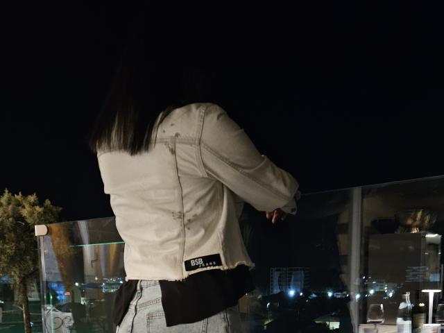 JoliDanielle staat met haar rug naar de camera, gekleed in een witte denimjas en lichtblauwe jeans, haar zwarte haar valt over haar schouders, in een omgeving met weinig licht..