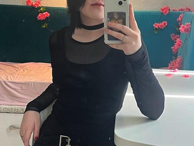EvitaCorolis in a black, sheer-top, long-sleeve, tight dress, taking a mirror selfie with a phone, visible nose and lips, short black hair, pink petals on white countertop.