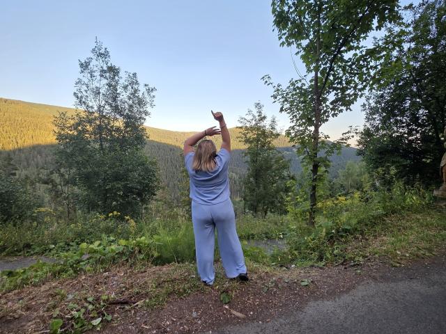 RadaRori stands with arms raised, back to camera, wearing blue pants and shirt, on a gravel path, forest and hills in background, blonde hair, green trees, clear sky.
