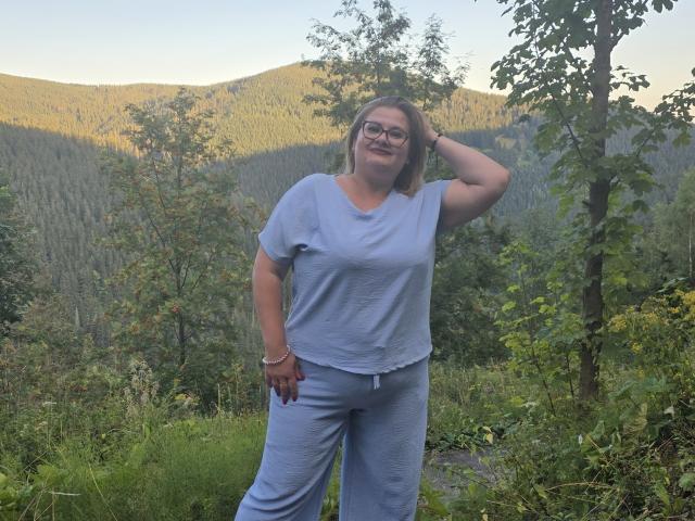 RadaRori stands in a forest, wearing a light blue shirt and pants, with glasses, medium tits, and a hand in her hair, smiling at the camera.
