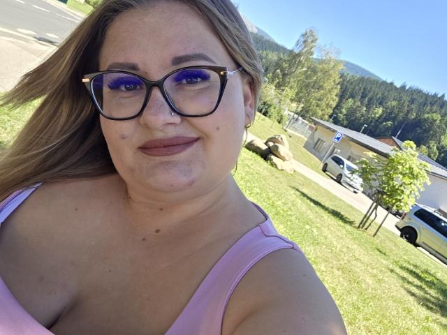 RadaRori with blue-tinted glasses, pink top, and freckles, smiling in a selfie, showing her chubby tits and nose ring, on a sunny grassy hill.