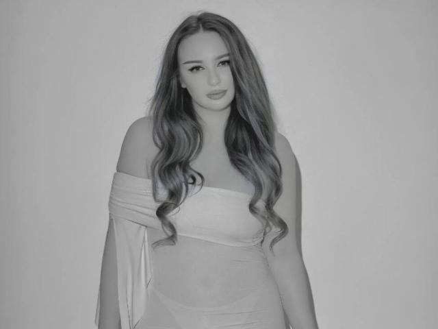 GitaOrtea has long, wavy hair, wearing a sheer, off-shoulder top that reveals her tits and a hint of her pussy. She's looking directly at the camera.
