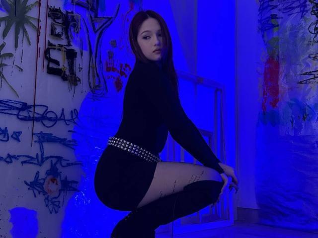 Bubochka, con cabello largo y castaño, una blusa negra de manga larga y un vestido negro corto con un cinturón con tachuelas, está en cuclillas, usando calcetines negros hasta la rodilla, en una habitación iluminada con luz azul, mirando a la cámara..