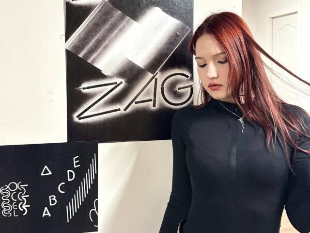 Bubochka, con cabello rojo, vestida con una camiseta negra ajustada de manga larga, mostrando sus pechos, de pie frente a una obra de arte en blanco y negro, mirando hacia abajo..