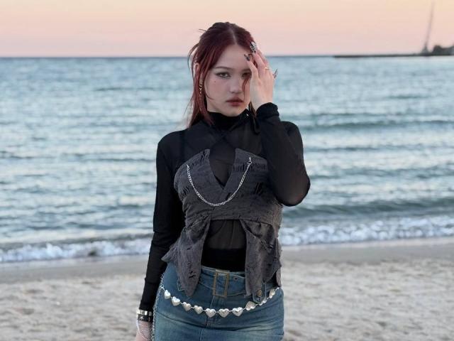Bubochka, com cabelo ruivo, blusa preta transparente, colete cinza texturizado, jeans azuis e cinto branco com formato de coração, em pé na praia, com a mão tocando a cabeça, o mar ao fundo, pôr do sol..