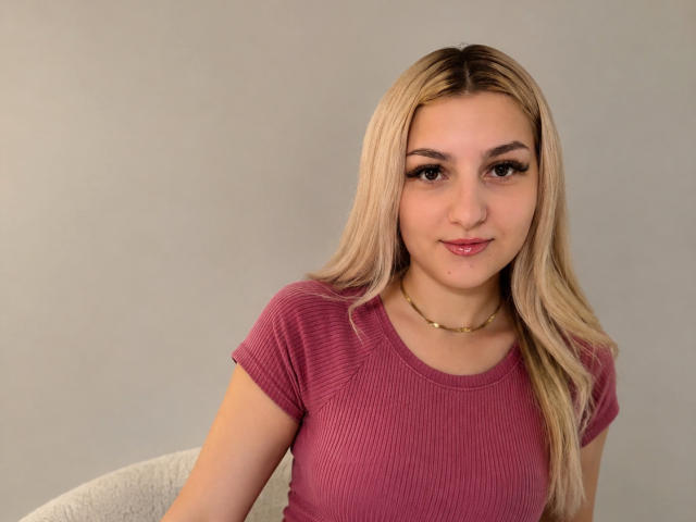 AngelofSinn, con i capelli biondi, una maglietta a coste rosa e una collana dorata, che mostra il seno e un sorriso appena accennato, seduta di fronte a uno sfondo neutro..