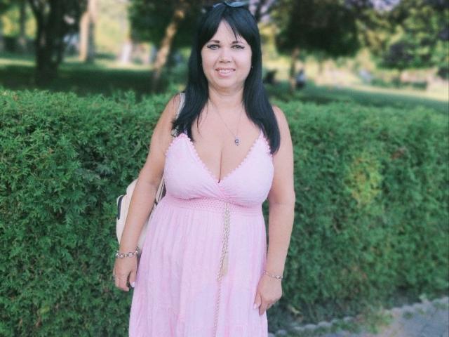MatureVivian has big boobs, black hair, and a pink dress with a deep V-neck, standing in front of a green hedge, smiling with a white bag on her arm.