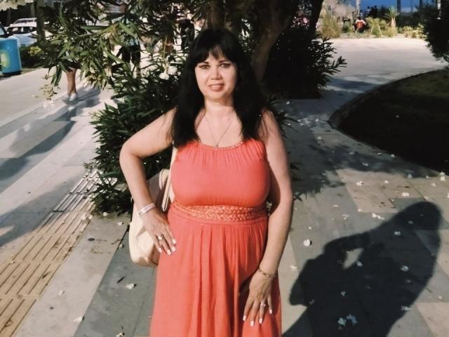 MatureVivian with black hair, orange dress, and white purse, standing on sidewalk, hands on hips, medium tits, shadow on ground, sunny day, leaves on pavement.