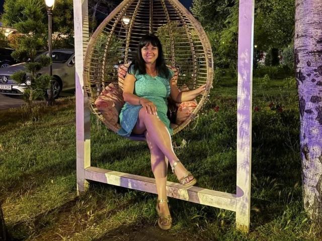 MatureVivian, with tits covered by a green dress, sits in a woven egg chair, holding a drink, legs crossed, wearing sandals, in a grassy, nighttime park setting.