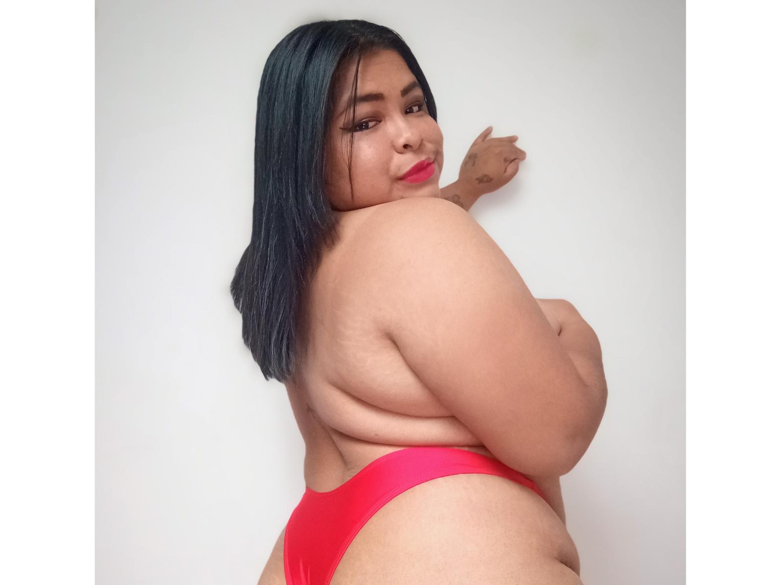 Zhory, con el pelo negro y oscuro, mirando hacia atrás, sin ropa en la parte superior, vistiendo ropa interior roja, mostrando su gran y curvilínea trasero, y su piel suave y morena. Está sonriendo, con una mano tocando la pared..