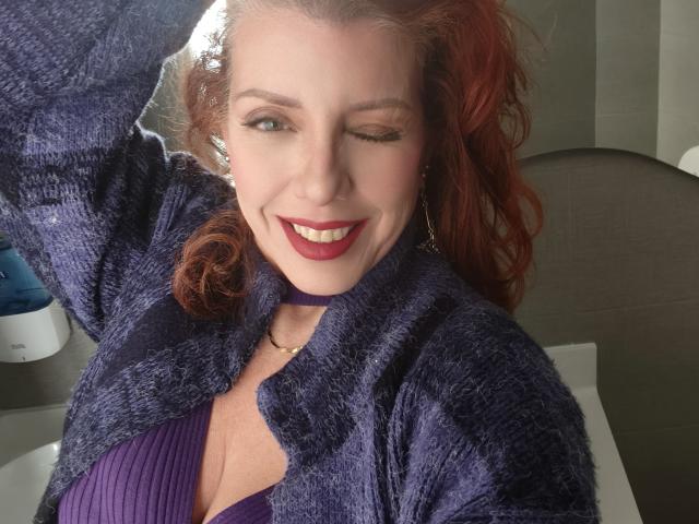 Khharha, con i capelli rossi, che fa l'occhiolino, sorridendo, indossa un cardigan viola peloso e un reggiseno viola, mostrando il seno, seduta su una sedia, con una mano tra i capelli..