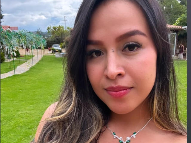 Het gezicht van LiaDumont, met lang donkerbruin haar, draagt een zilveren ketting met groene en witte stenen, ze glimlacht licht, op de achtergrond is groen gras en bomen te zien, en een bewolkte lucht..