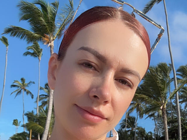Leahred's face, red hair pulled back, sunglasses on head, slight smirk, clear blue sky, palm trees in background, fair skin, subtle makeup, soft light, relaxed expression.