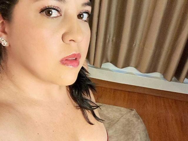 PaulaAnd tem uma pele clara, cabelo escuro preso em um rabo de cavalo, e está usando batom rosa com pequenas brincos de diamante, olhando para o lado com a boca ligeiramente aberta..
