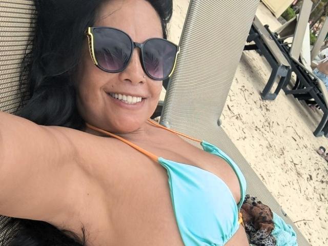 AntonellaMendoza lying on a chair, wearing a turquoise bikini top, orange straps, large tits, black sunglasses with yellow edges, smiling, tan skin, black hair, beach setting.