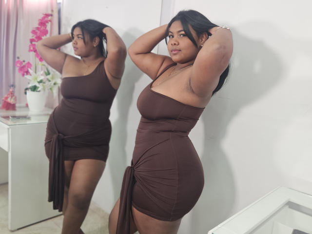 CameliaBloom est debout, vêtue d'une robe marron à une épaule avec un côté noué, les bras levés, reflétant sa silhouette aux courbes généreuses, sa peau brune, ses seins de taille moyenne dans un miroir..