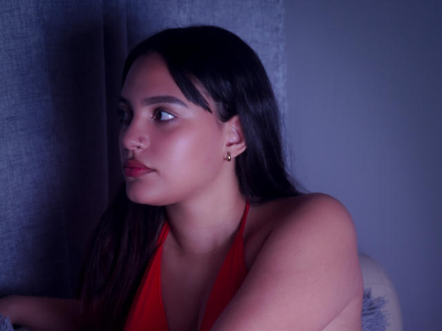 HeidyGloss con lunghi capelli neri, top rosso a fascia, piercing al naso e orecchini, che guarda di lato, illuminazione blu tenue e un leggero rossore sul viso..