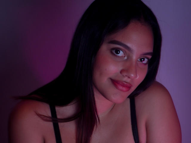 HeidyGloss with dark brown hair, nose ring, and pink lipstick, wearing a black strap top, smiling slightly, looking at camera, soft purple and pink lighting.