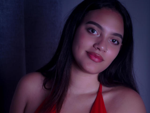 HeidyGloss met een medium bruine huid, zwart steil haar, rode lippenstift, een kleine neusring en een rode haltertop, die zachtjes naar de camera glimlacht, met blauw-grijze verlichting..