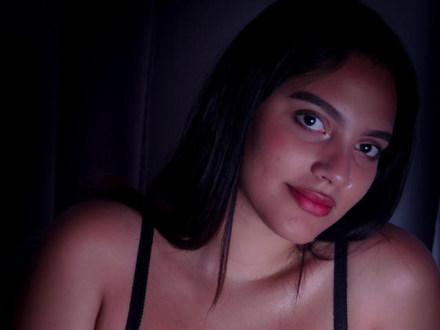 HeidyGloss has dark, straight hair, medium brown skin, and red lips, wearing a black spaghetti strap top, looking at the camera with a slight smile.