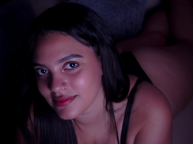 HeidyGloss, con i capelli scuri, un piercing al naso e le labbra rosse, sdraiata a pancia in giù, che mostra il sedere, indossa una maglietta nera e guarda la fotocamera con un sorriso appena accennato..