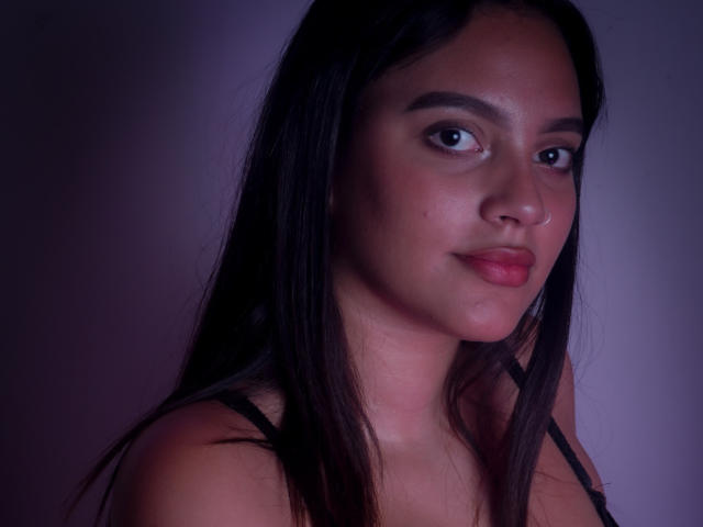 HeidyGloss hat langes, schwarzes Haar, braune Augen und einen Nasenring. Sie trägt ein schwarzes Oberteil, hat rosa-rote Lippen und ein leichtes Lächeln. Ihre Brüste sind nicht sichtbar..
