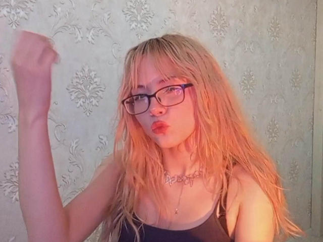 EileneSzala with wet blonde hair, glasses, pouty lips, black top, raising right fist, necklace, light skin, in front of ornate wallpaper. Boobs, ass, pussy not visible.