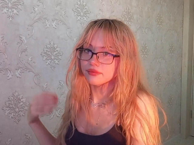 Blonde EileneSzala with wet, messy hair, black glasses, and a black top, showing off her small tits, raising her right arm, standing in front of a floral wallpaper.