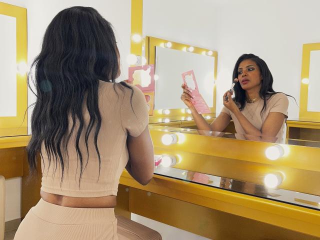 CamilaWest, con cabello largo, negro y ondulado, un top corto color beige y una falda a juego, está de pie frente a un espejo, mirándose a sí misma, sosteniendo un pincel de maquillaje rosa..