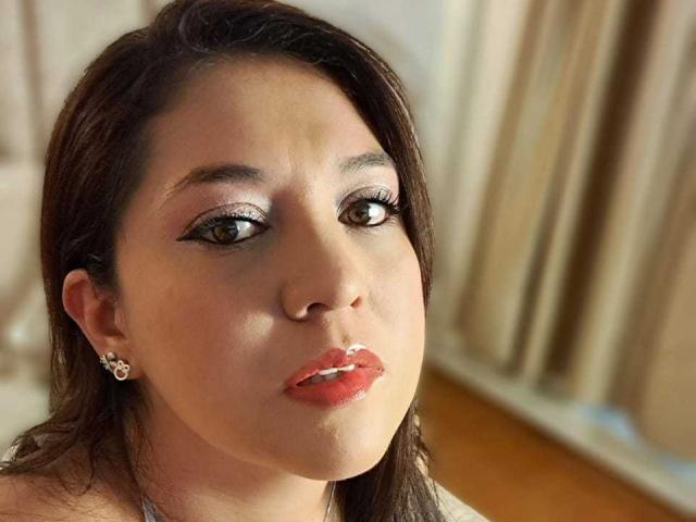 PaulaAnd tem olhos castanhos, cabelo castanho escuro e lábios vermelhos brilhantes, com um delineador discreto e brincos. Seu rosto é ligeiramente curvado. Ela está vestindo uma blusa branca..