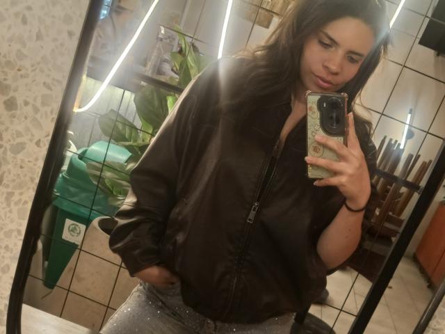 VioletaStone with wavy brown hair, wearing a black jacket and sparkly pants, taking a mirror selfie with hands in pockets, showing her cute face and small ass.