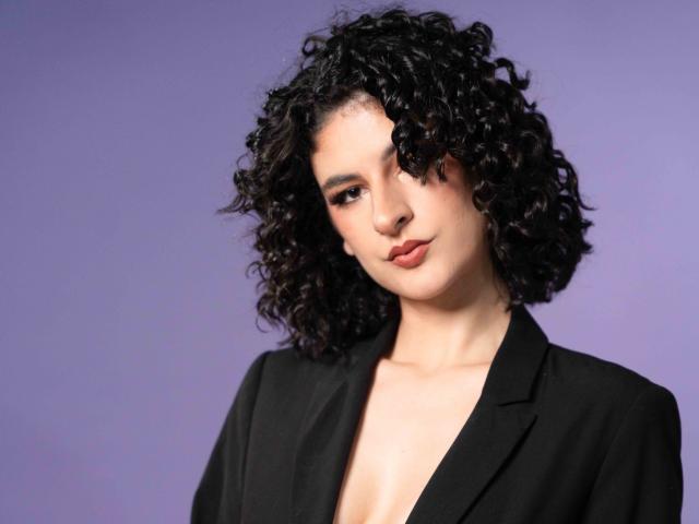 RoxyGraff with curly black hair, wearing a black blazer, has a slight pout and a confident, sexy look, showing a hint of her bare, small, perky tits.