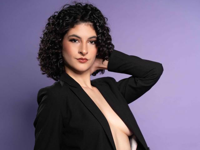RoxyGraff with curly black hair, wearing a black blazer, exposing her small bare tits, has a hand behind her head, against a purple background, looking confident.