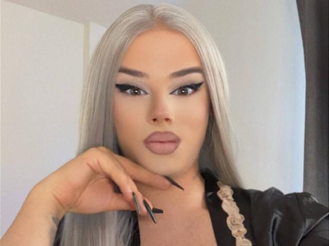 EvaLexy, com seus longos cabelos prateados, delineador preto marcante e lábios nude, veste uma jaqueta de couro preta, tem unhas pretas longas e uma tatuagem na mão..