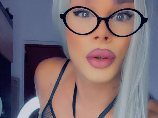 EvaLexy with blonde hair, wearing black glasses, has pink lipstick, visible tattoo on right shoulder, and is showing her tits in a black mesh top.