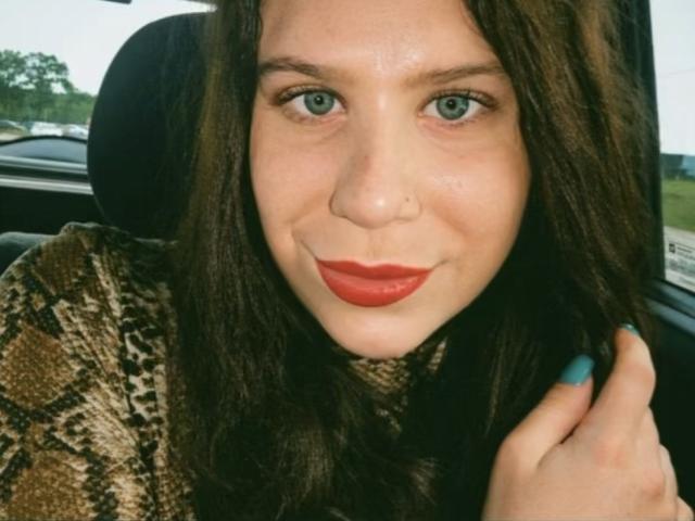 MayaFlowers tem olhos verdes, batom vermelho e esmalte azul. Ela está vestindo uma camisa com estampa de cobra, e seu cabelo castanho está solto. Ela está sorrindo levemente, segurando o cabelo..
