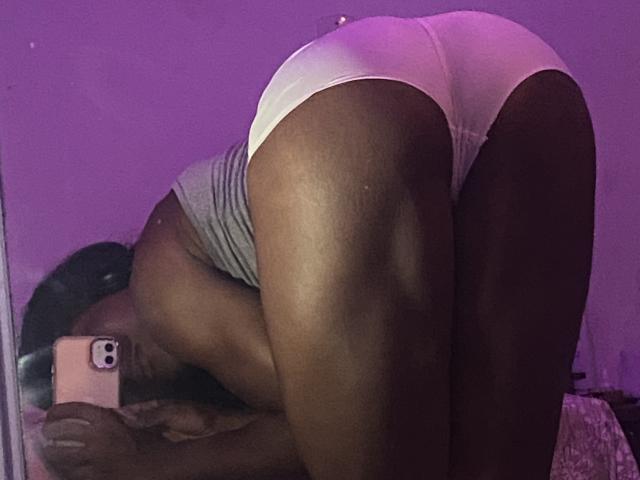 PretinHatszl bending over, showing a dark-skinned, muscular ass in white panties, wearing a gray tank top, taking a selfie with a pink iPhone in a purple-lit room.