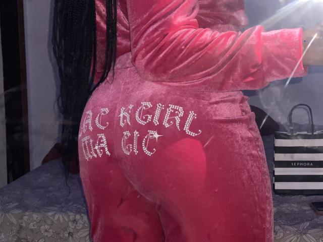 PretinHatszl in a pink velvet onesie with "BLACK GIRL" in sparkly letters across her butt, standing with her back to the camera, long black braids, room with shopping bags.