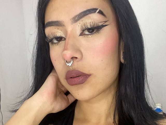 QueenMissty a de longs cheveux noirs, un anneau de nez en forme de papillon, et un eye-liner noir épais avec des ailes spectaculaires. Ses joues roses et ses lèvres brunes brillantes contrastent avec sa peau bronzée..