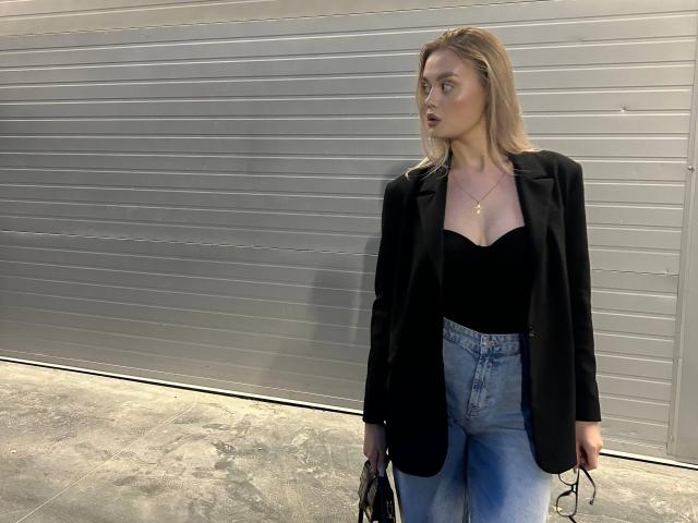 OliviaAzbill has blonde hair, black blazer, black strapless top, blue high-waisted jeans, holding black handbag, looking to the side with slightly open mouth.