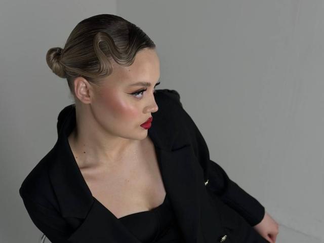 OliviaAzbill has wavy brown hair in a bun, red lipstick, and a black outfit with a shiny collar, looking to the side with blue eyes and pink blush.