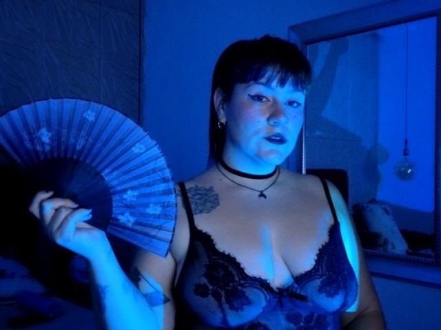 NaniSky with blue-tinted skin, black lace bra showing tits, holding a fan, choker necklace, tattoo on right shoulder, short black hair, looking at camera, dark lipstick.