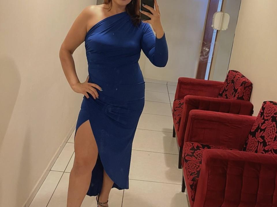 MorenaOn in a blue, one-shoulder dress with a high slit, showing her leg and black heels, standing in a room with red, patterned chairs.
