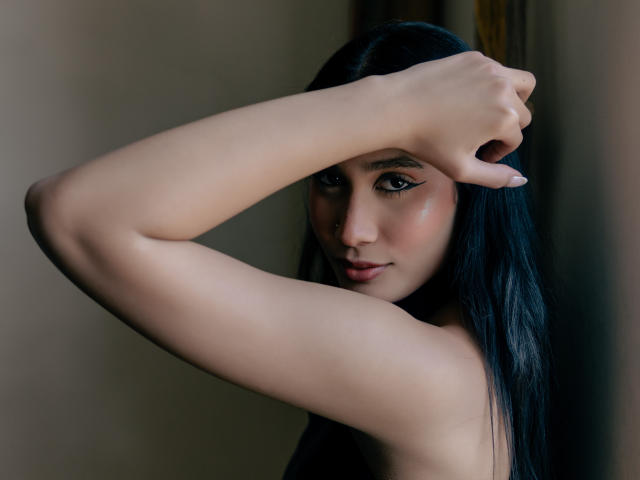 JennTylor with long black hair, arms raised, covering one eye, showing off her cute nose, subtle makeup, and smooth skin, looking seductively at the camera.
