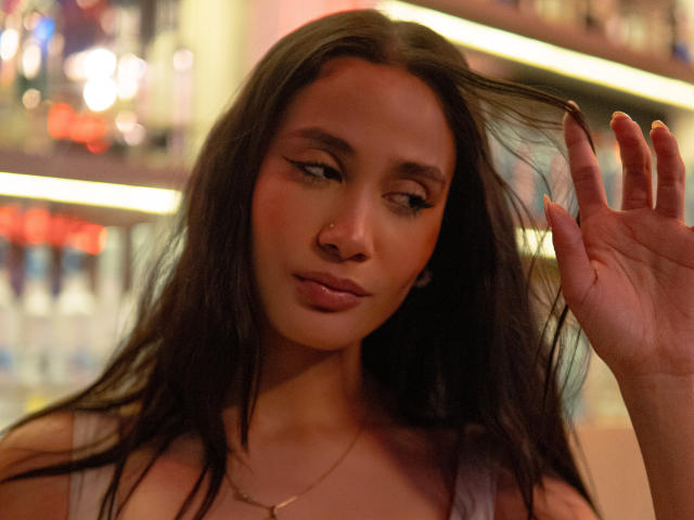 JennTylor with long dark hair, slightly parted lips, and a small nose piercing, holding hair with a hand, wearing a sleeveless top, in a warmly lit bar.