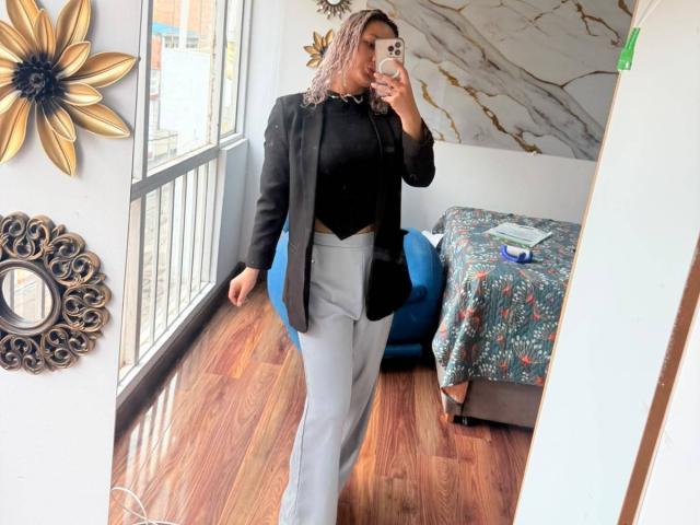 Elena Saenz mit welligem, rosa Haar, schwarzem Oberteil, schwarzem Blazer, weißen Hosen, einer schwarzen Umhängetasche, die ein Spiegel-Selfie macht, auf einem Holzboden steht und ein iPhone hält..