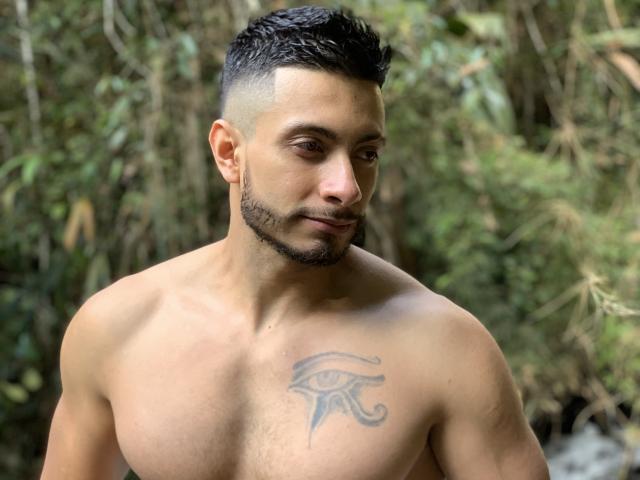 Markbuffet01, senza camicia e con un tatuaggio di un occhio sul petto destro, ha un taglio di capelli corto e scuro, quasi rasato, e una leggera barba, ed è in piedi in una foresta..