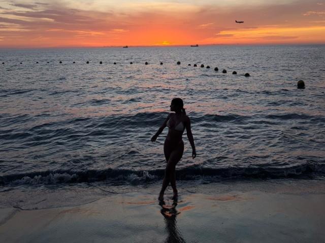 Silhouetted ThaliaAres with wavy hair, small tits, and a slim ass, walks on a wet beach at sunset, ocean waves gently touching her legs. A bird flies overhead.