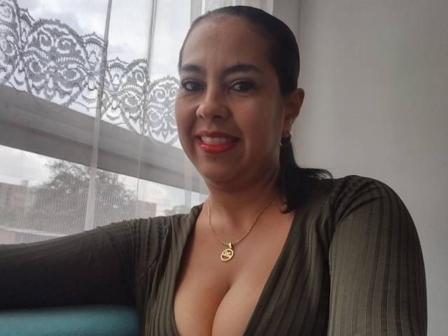 Kathiusca with dark hair, red lipstick, green dress, deep cleavage, gold necklace, smiling, standing by a window with lace curtain. Her tits are visible.