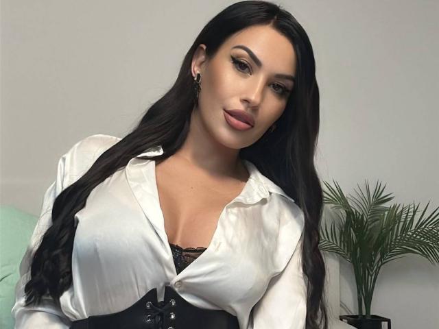 ZoeyMarie with long black hair, wearing a white satin blouse, black lace bra, and black corset, showing her big tits, and a subtle smile.
